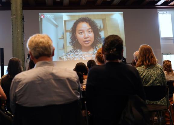 sitzende Personen von hinten aufgenommen während sie einer online Keynote auf einer Leinwand zuschauen.